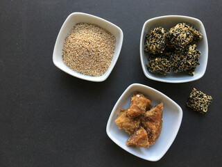 Organic jaggery, raw white sesame seeds and sesame bar displayed in various bowls. Preparation for homemade sesame energy bars. View from above on black background.