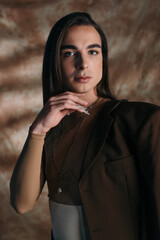 Naklejka premium Portrait of stylish nonbinary person touching chin and looking at camera on abstract brown background.