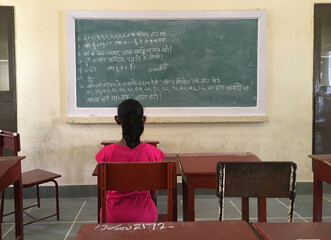 A young girl student looking at a blackboard with in the classroom. A back view of a pupil in the classroom of an Indian school.