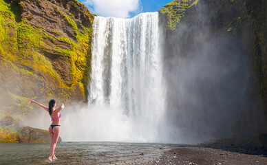 Beautiful girl view from back in bikini raising arms in front of waterfall - Icelandic Landscape concept - View of famous Skogafoss waterfall 