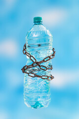 Plastic water bottle tied with rusty chains on blue skyes background.