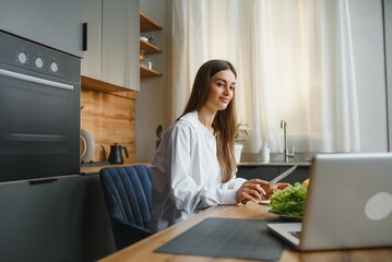 Athletic woman blogger nutritionist prepare a salad with fresh vegetables and conducts a video conference on healthy eating on laptop in the kitchen