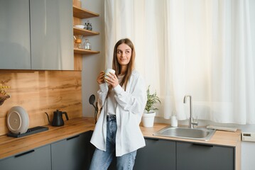 A young beautiful caucasian woman stands in the kitchen with a white cup of coffee or tea in the morning. A lonely girl is enjoying a cup of fresh hot drink.