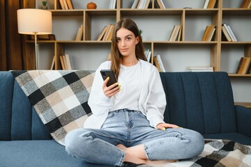 Smiling young Caucasian woman sit relax on couch in living room talk on video call. Happy millennial female rest on sofa at home have webcam digital virtual communication on cellphone.