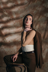 Nonbinary person in corset holding jacket near chair on abstract brown background.