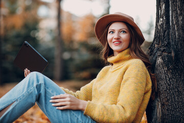 Young woman model sitting with digital tablet in autumn park with yellow foliage maple leaves. Fall season fashion