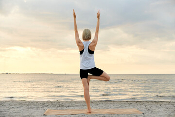 Fototapeta na wymiar fitness, sport, and healthy lifestyle concept - woman doing yoga tree pose on beach over sunset