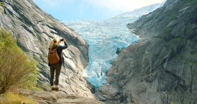 Global warming, melting glaciers and rising sea levels on rocky mountains view in nature. Back view of ecologist doing research outdoors. Woman looking at a landscape and taking photos with a phone