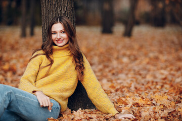 Young woman model sitting in autumn park with yellow foliage maple leaves. Fall season fashion