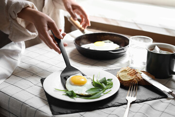 Woman putting tasty fried eggs onto plate at table indoors, closeup