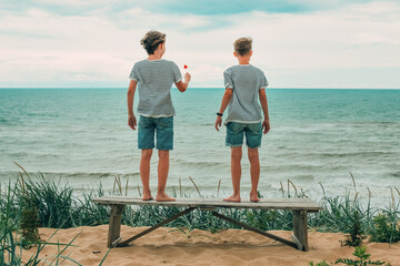 Two brothers are standing on a bench on a high sandy seashore in the wind, looking into the distance, a flower in their hand