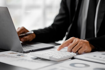 Obraz premium Businessman is using a calculator to calculate company financial figures from earnings papers, a businessman sitting in his office where the company financial chart is placed. Banner with copy space.