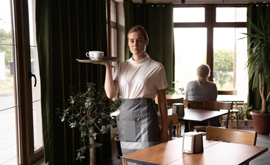 Portrait of cheerful waitress serving on tray cake and cup of beverage in cafe
