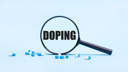 On a blue background, white and blue capsules with pills and a black magnifying glass with the text DOPING. Medical concept.