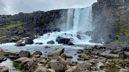 Blue waterfall against black rock mountains with green moss under cloudy sky in Iceland