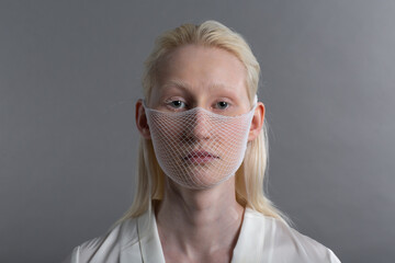 Closeup portrait of white caucasian albino blond woman fashion model wearing quarantine medical face mask coarse mesh net on grey background. Covid-19 coronavirus protection concept. - obrazy, fototapety, plakaty
