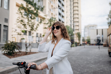 Fototapeta na wymiar Beautiful young woman in sunglasses and white suit standing on her electric scooter near modern building and looking away
