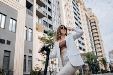 Beautiful young woman in sunglasses and white suit standing on her electric scooter near modern building and looking away