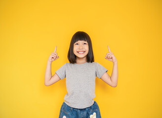 Portrait of young fun smart happy little cute asian girl isolated on yellow with copy space studio shot. Education for elementary kindergarten, little girl finger point up back to school concept