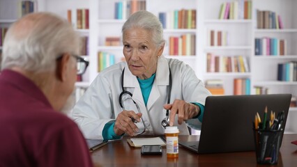 Serious old senior female general practitioner in lab coat holding digital computer tablet, showing laboratory test results to interested elderly 70s male patient at checkup meeting in hospital.