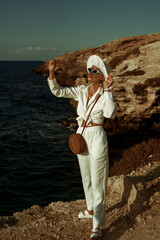 Elegant fashionable woman wearing white linen suit, straw hat, sunglasses, leather sandals, with round wicker crossbody bag, posing on rocks near the sea. Outdoor summer fashion full-length  portrait 