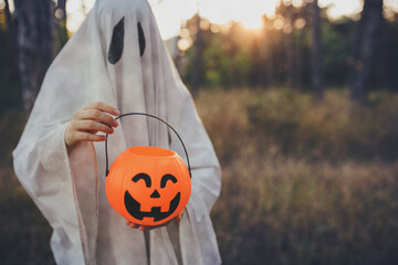 Girl wearing ghost costume holding pumpkin bucket with candies, standing in a forest.
