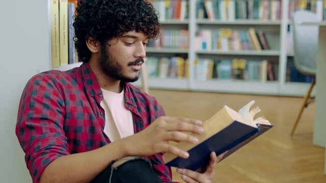 Book reading. Handsome clever indian or arabian university or college male student, sitting on a floor in a library near bookshelf, reading interesting book, preparing for exam, gain knowledge, smile