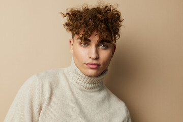 Obraz premium portrait of a handsome man, with curly hair, standing on a beige background in a light turtleneck, looking pleasantly into the camera. A photo with an empty space for inserting an advertising layout