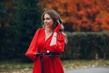 Young woman with electric scooter in red dress at the autumn city park.