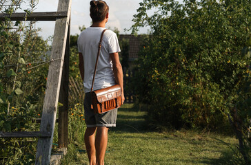 young man wearing a small vintage leather bag handbag over his shoulder standing outside in the...