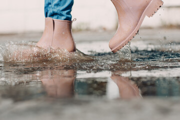 Woman wearing rain rubber boots walking running and jumping into puddle with water splash and drops in autumn rain.