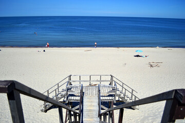 Beach on the Curonian Spit (Kurshskaya Kosa). Almost white sand. Blue calm Baltic Sea. In the foreground is a wooden staircase descending to the beach. Symmetrical horizontal photo, Kaliningrad region