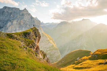 A fearless hiker stands on an overhanging rock and enjoys the view mountain landscape of the Alps in summer with the sun rising over the mountain peaks in the distance.
