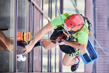 Industrial mountaineering worker washing glass windows of high-rise building, hanging on safety...