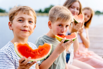Four children boys and girls sitting on the row on wooden bridge by the lake eating watermellonn fresh juice friuts. Outdoor travel vacation healthy lifestyle vegan food picnic hungry children with sn
