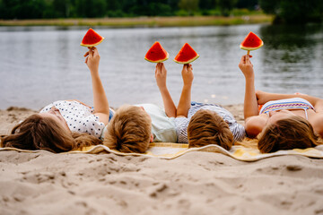 Group of four different age children eating watermelon lying on beach. Two girls and two boys friends holding slice of watermelon on stick enjoy summer together. Friendship, good mood, leisure.