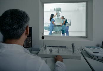 Female nurse preparing mature patient for X-ray of hand near X-ray machine while the radiologist waits behind protective window in office