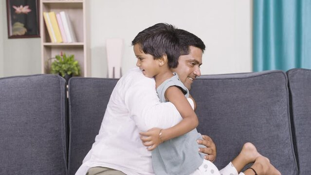 playful father kissing by hugging little his daughter while sitting on sofa at home - concept of family time, fatherhood and positive emotion.