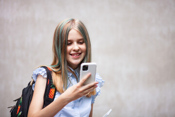 Teen schoolgirl with backpack smiling and looking in smartphone on a grey wall background. Teens and mobile phones concept. Selective focus.