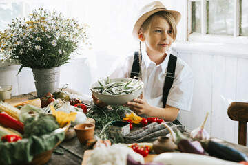 cute farmer boy in casual clothes sit near table on veranda of country village house with different vegetables, eco-friendly food from garden, autumn harvesting