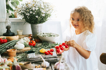 cute little girl in casual white dress sit near table on veranda of country village house with different vegetables, eco-friendly food from garden, autumn harvesting