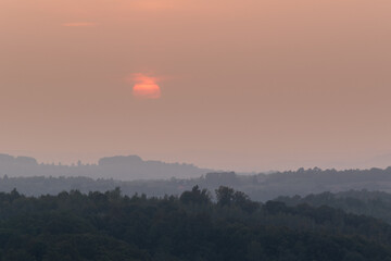 Atmospheric landscape scene of lush forest layers in mist during autumn sunset in countryside, forest obscured in fog and big orange sun dimmed behind clouds