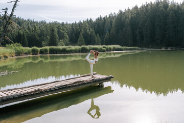 Eine Junge Frau praktiziert Yoga auf einem Steg am See zum Sonnenaufgang. Der Felixer Weiher - Tretsee in Südtirol. Weiße Leggins 2