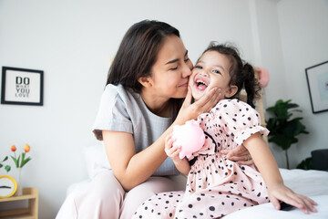 Happy cute Asian little daughter smiling and tender kiss from mom, sitting on bed, speaking,...