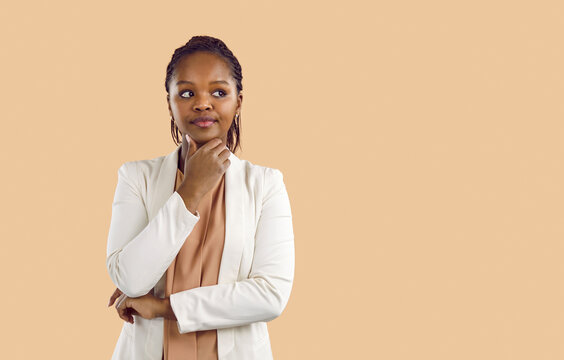 Portrait of young black business woman holding hand on chin, dreaming, imagining, thinking about something and looking away at blank empty copyspace on right side of solid beige background