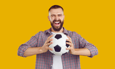 Funny, crazy male football fan supporting his team. Portrait of a happy, excited young man in a casual shirt standing isolated on a yellow color background, holding a soccer ball and screaming loudly