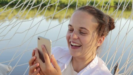 Dark haired smiling positive optimistic female with bun hairstyle hanging in hammock near the river and holding smart phone in hands, having video call, waving hand, saying hello.