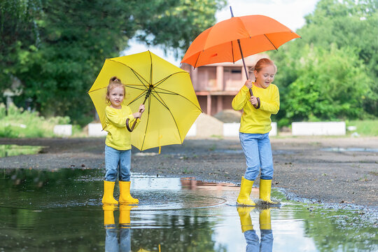 two little girls sisters walk in warm weather after rain through puddles in rubber boots and under bright yellow umbrellas