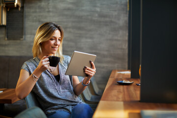 Young beautiful woman relaxing in coffee shop reading electronic book via internet on digital tablet and drink coffee
