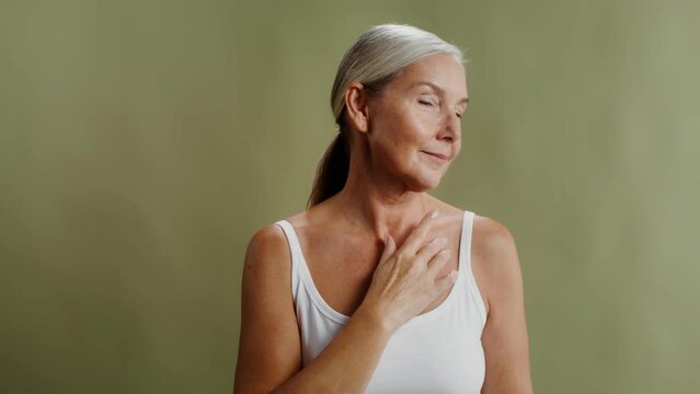 Elderly gray-haired model posing while looking at the camera and touching her face and d&eacute;collet&eacute; with her hands while standing against a monochrome studio background